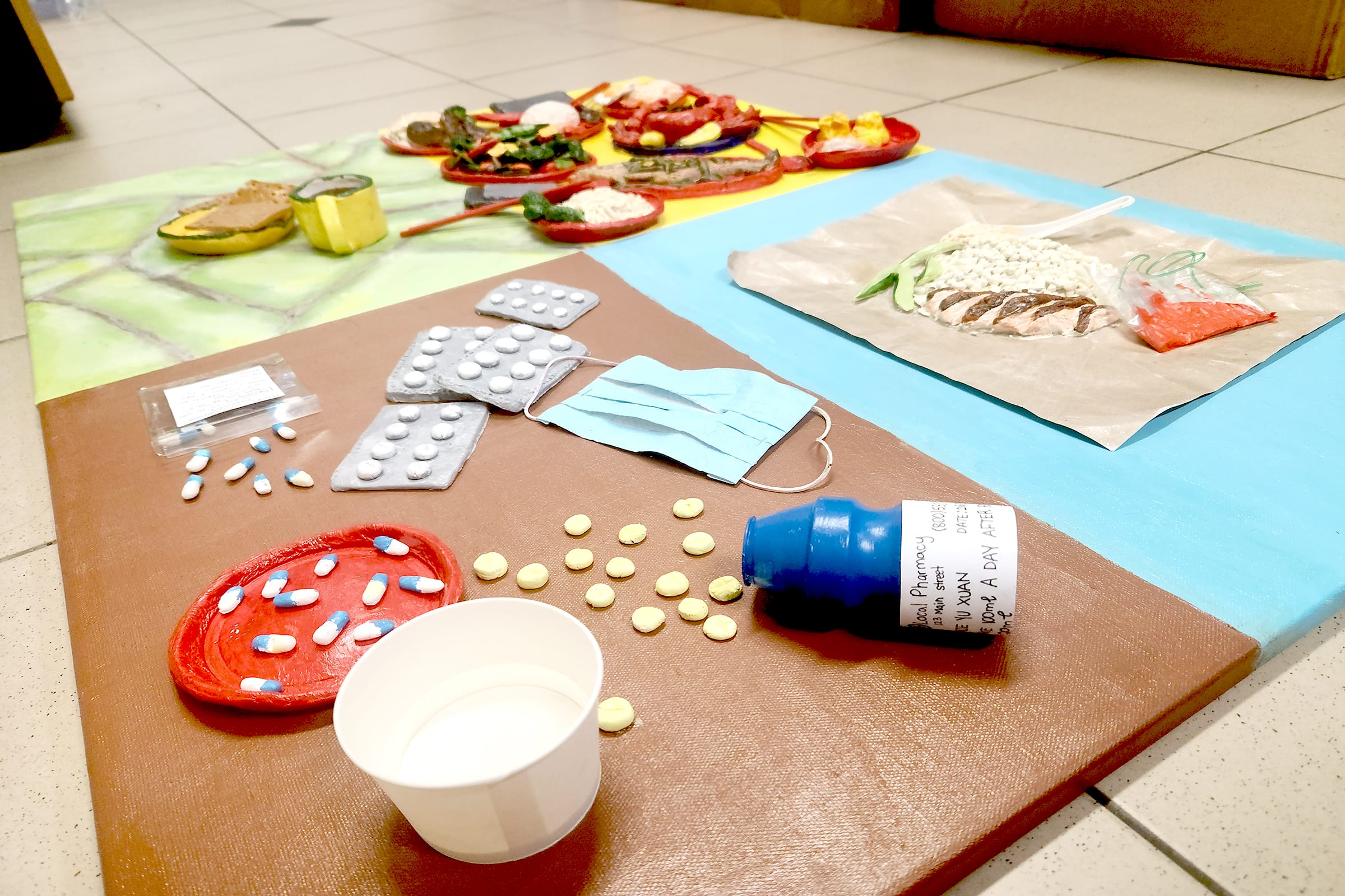 Handmade clay models of pills, pill packs, a face mask, medicine bottle, and food displayed on colored boards.